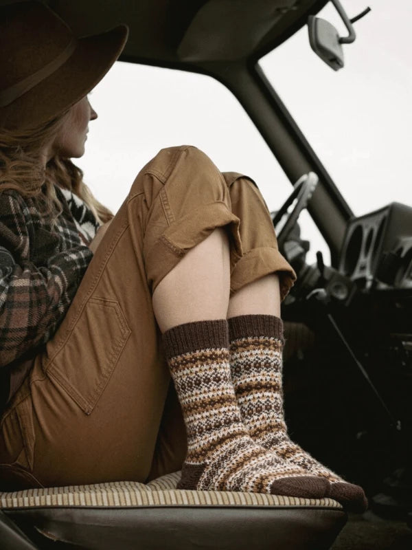 A person wearing brown patterned wool socks—perhaps crafted from Ronja Hakalehto’s Kiertoteitä—rolled-up tan pants, and a plaid shirt sits in the driver’s seat of a vehicle, legs on the seat, gazing out at Pohjois-Suomi.
