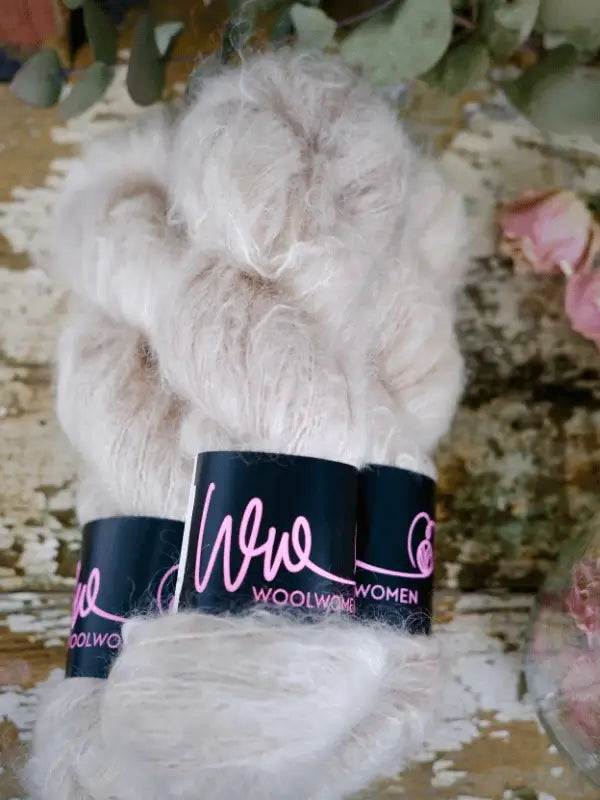 Three skeins of WoolWomen Fluff Mohair in creamy käsinvärjätty yarn, labeled in pink script, are displayed on a rustic surface with dried flowers nearby.