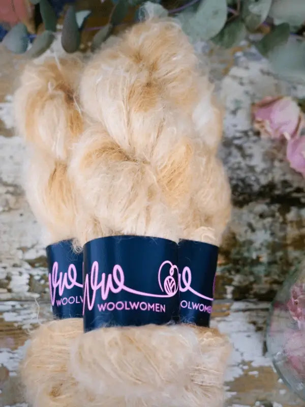 Three skeins of golden-yellow WoolWomen Fluff Mohair with a pink logo, beside flowers and greenery—perfect for your next project using the Fussa neuletakki neuleohje + WoolWomen Fluff Mohair set.
