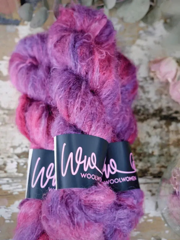 Two skeins of WoolWomen Fluff Mohair yarn in pink and purple, bundled with WOOLWOMEN labels, are displayed on a rustic wooden surface—ideal for your cozy projects.