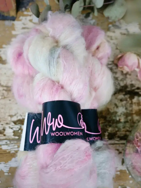 A skein of WoolWomen Fluff Mohair in pale pink and white, wrapped with a black label, rests on a rustic wooden surface. This käsinvärjätty mohairlanka is perfect for cozy projects.