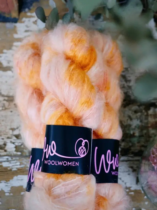 A close-up of two skeins of WoolWomen Fluff Mohair yarn in orange and light pink on a rustic wooden surface, with blurred greenery in the background.