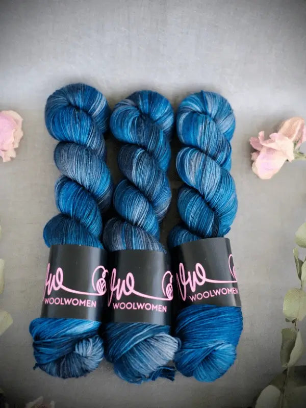 Three skeins of blue and gray variegated WoolWomen Merino Sock yarn are lined up side by side, each with a pink label. Light pink flowers and green leaves frame the image on a gray background.