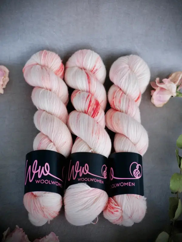Three skeins of WoolWomen Merino Sock, soft pastel pink and white käsinvärjätty lanka with subtle red streaks, each wrapped in a black label, are displayed on a grey background with delicate flowers.