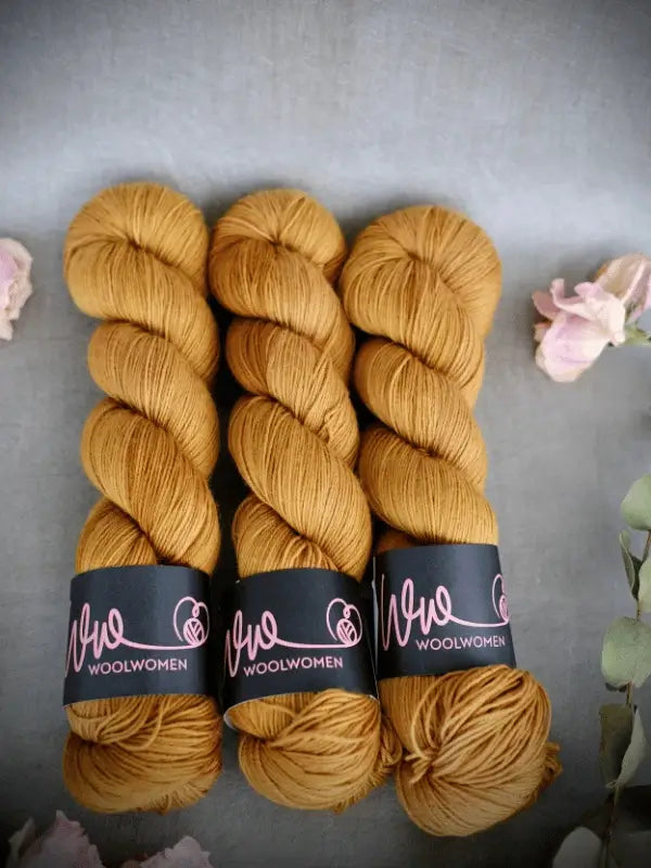 Three skeins of golden yellow WoolWomen Merino Sock yarn are neatly placed side by side on a gray surface, each labeled. Soft pink and green floral accents decorate the corners of the image.