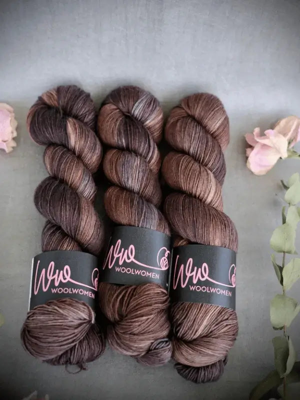 Three skeins of WoolWomen Merino Sock yarn in brown and gray variegated shades, made from käsinvärjätty lanka, are neatly arranged on a gray surface with light pink flowers and green foliage.