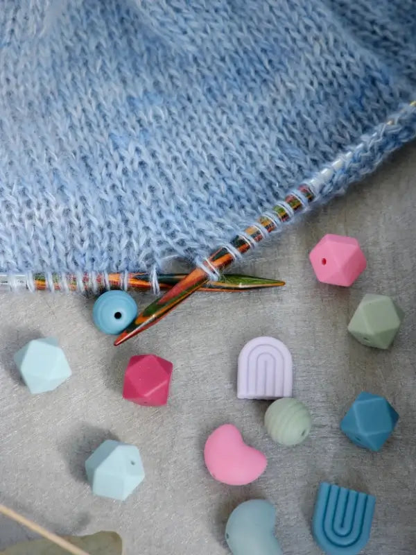 Close-up of blue knitted fabric on circular neulepuikot, with colorful geometric and arch-shaped Woolwomen Puikkostopparit and stitch markers scattered on a gray surface.