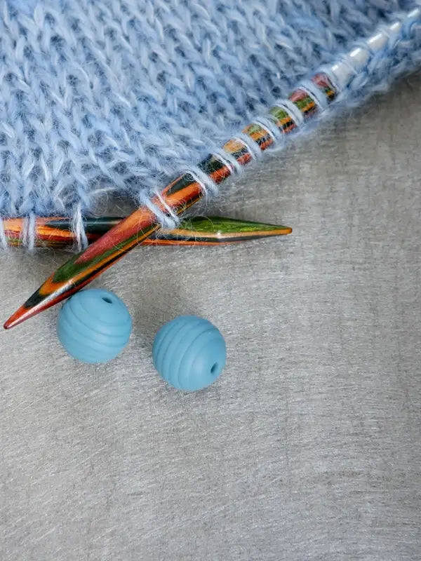 Woolwomen Puikkostopparit colorful wooden knitting needles with blue yarn in progress, accompanied by two round blue Woolwomen Puikkostopparit bead stoppers, all arranged on a gray textured surface.