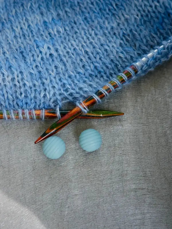 A close-up of blue knitted fabric on multicolored knitting needles featuring Woolwomen Puikkostopparit, with two light blue round buttons resting nearby on a gray surface.