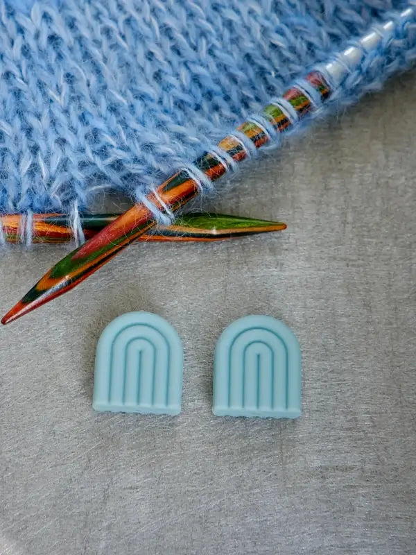Two colorful wooden knitting needles with light blue yarn and a pair of Woolwomen Puikkostopparit—light blue, arch-shaped stitch markers—rest on a gray surface.