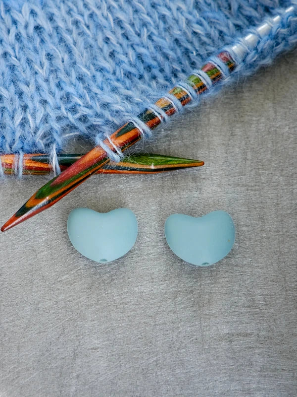 Two blue, heart-shaped Woolwomen Puikkostopparit rest on a gray surface alongside two colorful wooden knitting needles and a piece of blue knitted fabric.