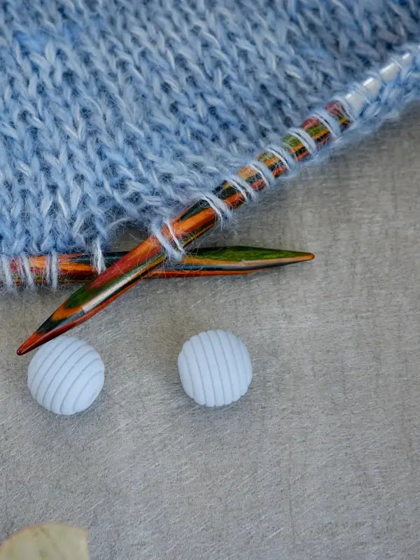 Sure! Here’s a revised version of your sentence using the product name: Colorful wooden knitting needles inserted into light blue yarn, with two Woolwomen Puikkostopparit—white, ribbed, round button-like accessories—resting nearby on a gray surface.