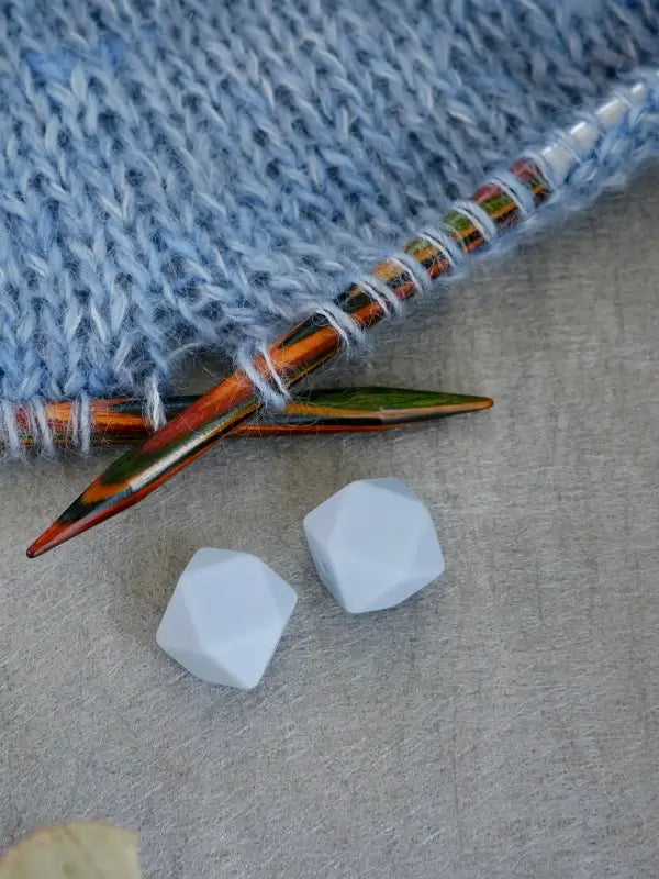 Close-up of Woolwomen Puikkostopparit colorful wooden knitting needles with light blue yarn, two white geometric beads, and a beige surface underneath.