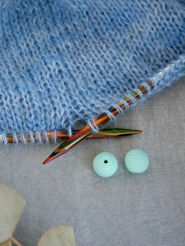 A close-up of blue knitted fabric on wooden knitting needles, accompanied by two Woolwomen Puikkostopparit in light blue, resting nearby on a gray surface.