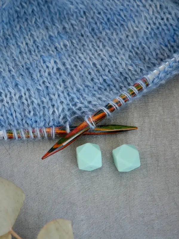 A close-up of light blue knitting on multicolored wooden needles, with two faceted mint-green Woolwomen Puikkostopparit lying on a gray surface nearby.