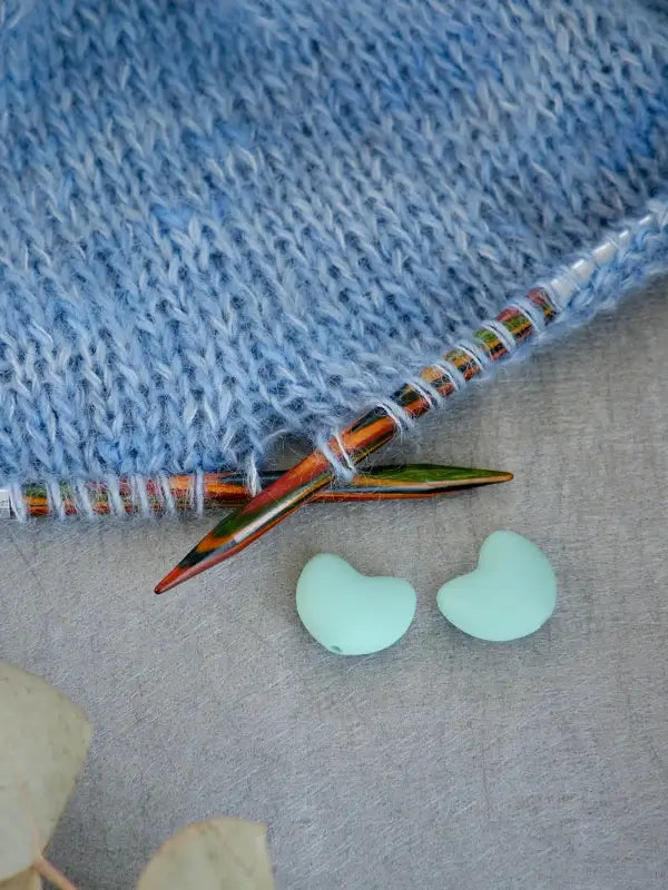 A close-up of a blue knitted fabric on colorful knitting needles, with two mint-green Woolwomen Puikkostopparit heart-shaped buttons placed nearby on a gray surface.