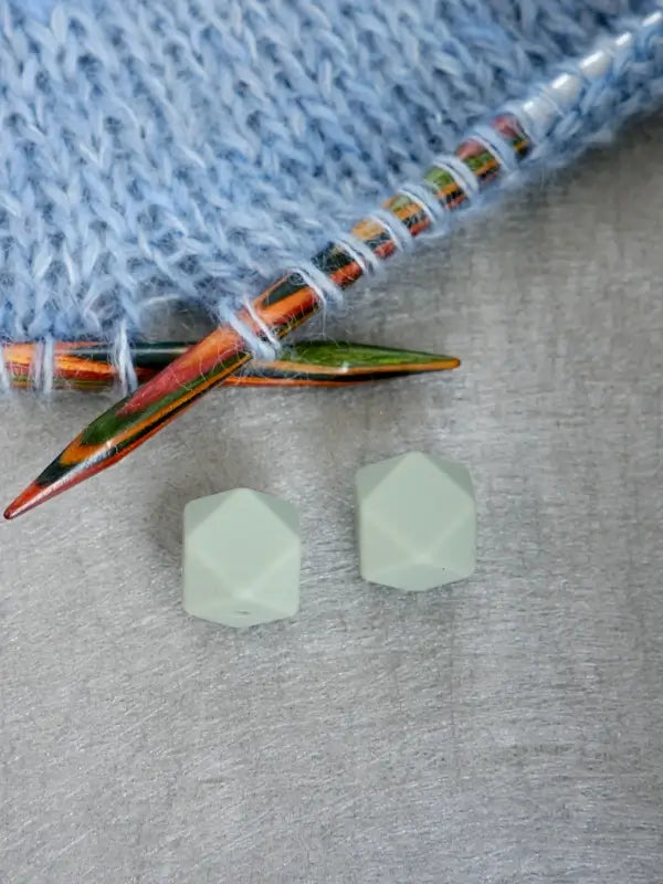 Two pale green, faceted beads rest on a gray surface near Woolwomen Puikkostopparit knitting needles and light blue knitted fabric.