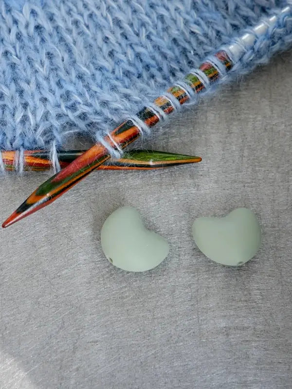 Two mint green Woolwomen Puikkostopparit, bean-shaped stitch markers, rest on a gray surface beside colorful wooden knitting needles and light blue knitted fabric.