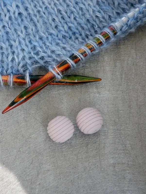 Two colorful Woolwomen Puikkostopparit knitting needles and light blue yarn rest near two round, pale pink buttons with textured lines, all placed on a gray fabric background.