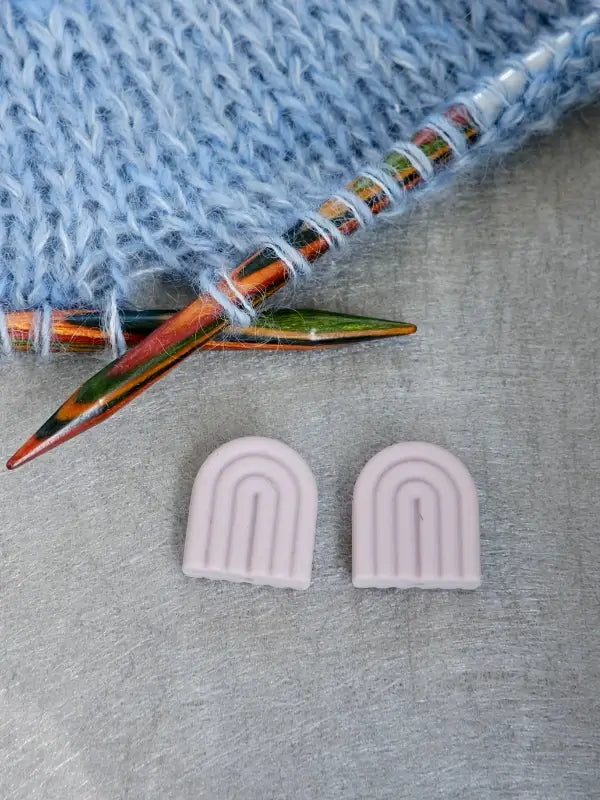 Two colorful knitting needles with blue yarn are next to two small, light purple, arch-shaped Woolwomen Puikkostopparit on a gray surface.