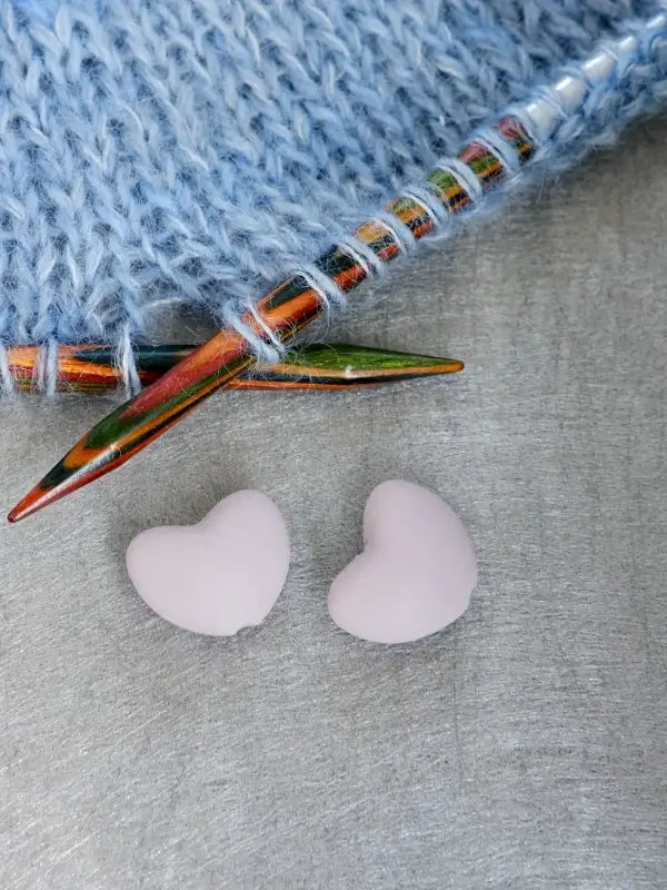 Two light pink Woolwomen Puikkostopparit, heart-shaped knitting needle stoppers, rest on a gray surface beside blue knitted fabric and colorful wooden knitting needles.