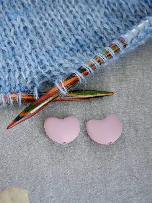 Two pink, bean-shaped Woolwomen Puikkostopparit rest on a gray surface next to colorful knitting needles and a swatch of light blue knitted fabric.