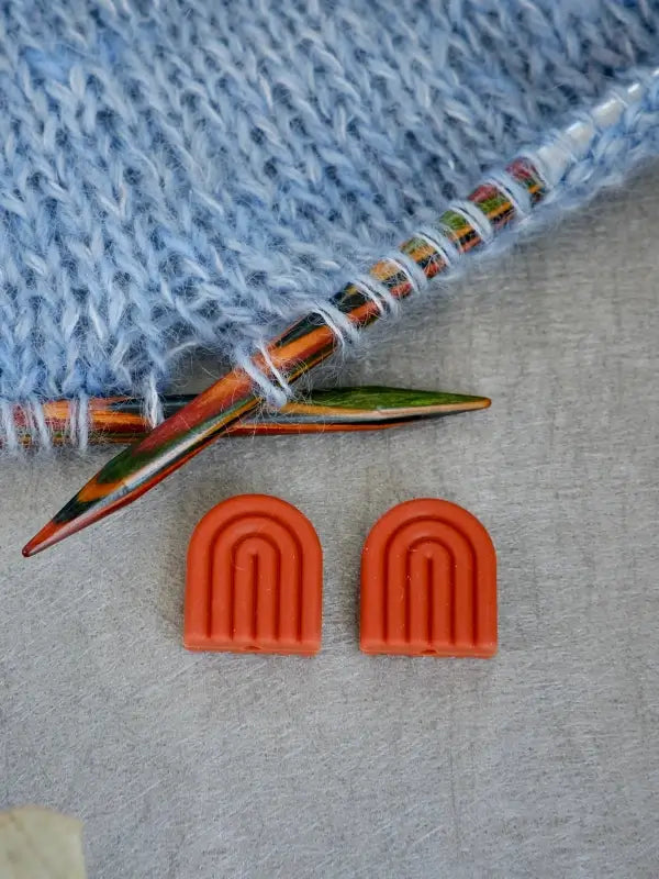Two orange Woolwomen Puikkostopparit, shaped like rainbows, rest on a gray surface next to colorful wooden knitting needles and light blue knitting in progress.