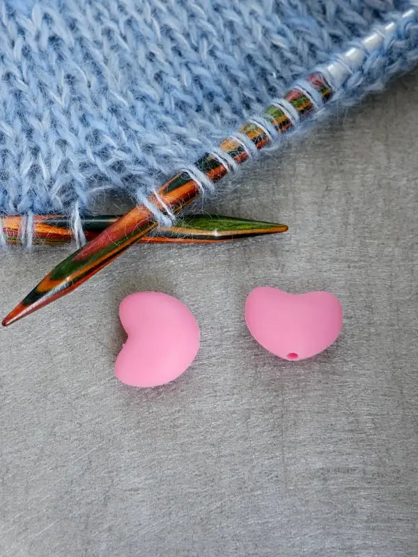 Two pink, heart-shaped Woolwomen Puikkostopparit rest on a gray surface beside colorful wooden knitting needles and blue knitted fabric.