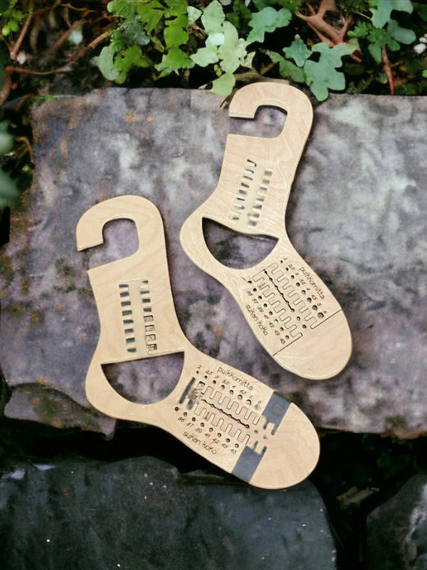 Two WoolWomen säädettävä sukkaplokki, crafted from kotimainen koivuvaneri with engraved measurements and hooks on top, rest on stones among green plants—perfect for shaping villasukat.