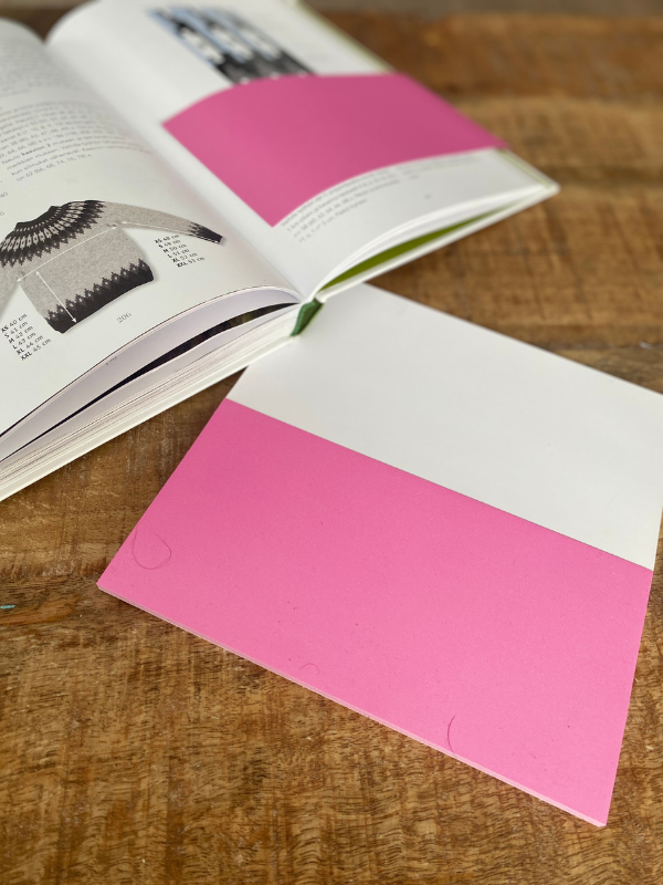 A book with knitting patterns lies open on a wooden table next to a piece of Staattinen paperi, pinkki—perfect for neulekaavion seuraaminen or making muistilaput as you work.
