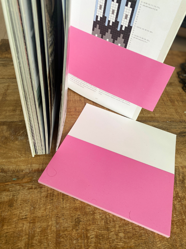 A Staattinen paperi, pinkki sheet, divided into pink and white sections—ideal for neulekaavion seuraaminen—lies flat on a wooden table next to an open magazine, with another sheet tucked inside.